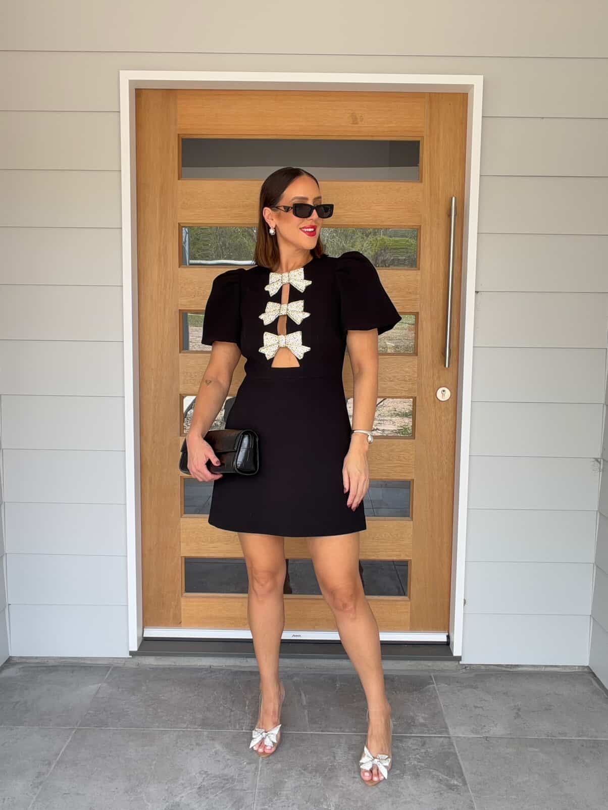 Woman in a black dress with white bow details standing in front of a wooden door.